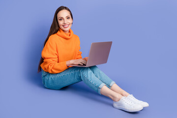 Full size body photo of cheerful young lady wear knitted orange pullover work in coworking with laptop isolated on purple color background