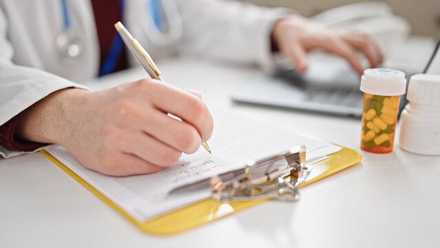 Young hispanic man doctor writing on clipboard using laptop at clinic