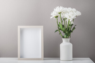 Empty photo frame and a bouquet of white chrysanthemums, daisies in a ceramic white vase. Postcard, festive home interior. Mockup.