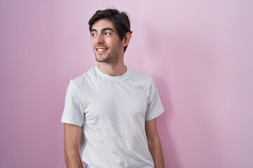 Young hispanic man standing over pink background looking away to side with smile on face, natural expression. laughing confident.