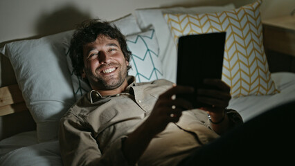 Young hispanic man using touchpad lying on bed smiling at bedroom