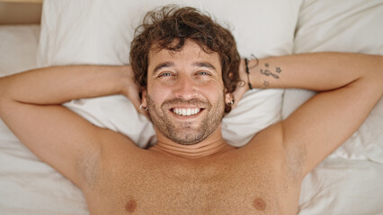 Young hispanic man relaxed on bed smiling shirtless at bedroom