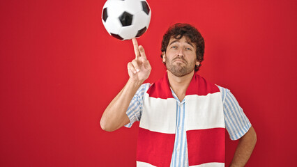 Young hispanic man supporting soccer team playing with ball over isolated red background