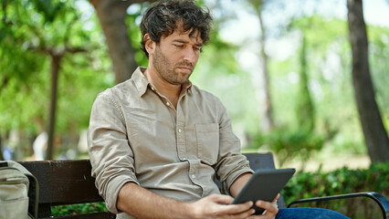 Young hispanic man using touchpad sitting on bench at park