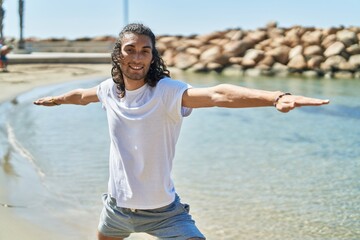 Young hispanic man doing yoga exercise standing at beach