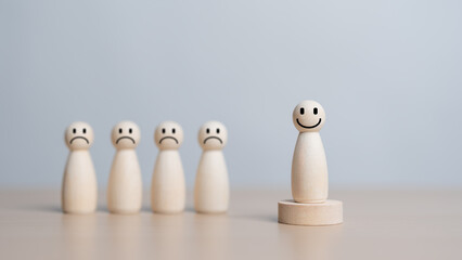 Smiley happy face wooden figure stands isolated from the sad crowd on a wooden table. Emotion...