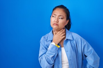 Asian young woman standing over blue background touching painful neck, sore throat for flu, clod...