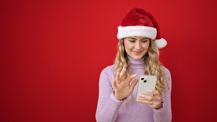 Young blonde woman wearing christmas hat using smartphone over isolated red background