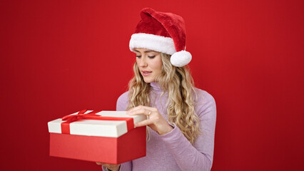 Young blonde woman wearing christmas hat unpacking gift looking upset over isolated red background