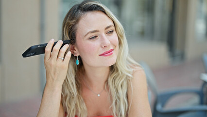 Fototapeta premium Young blonde woman listening to voice message sitting on table at coffee shop terrace