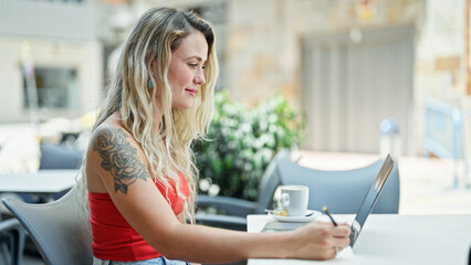 Young blonde woman using laptop taking notes smiling at coffee shop terrace