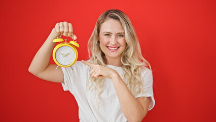 Young blonde woman smiling confident pointing to alarm clock over isolated red background