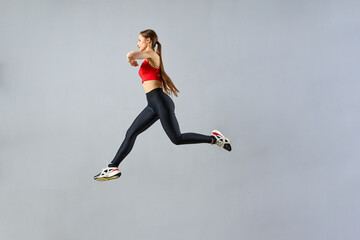Young fitness lady jumps high into the air on a gray background. A female trainer in sportswear performing a cardio workout. - obrazy, fototapety, plakaty