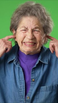 Vertical video of displeased irritated frustrated sad angry elderly senior woman with wrinkled skin closed eyes cover ears do not want to listen scream on green screen background studio portrait