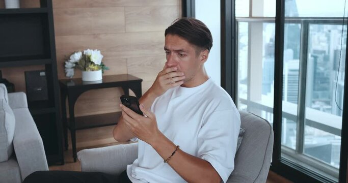 Young man receives notification with bad news on phone. Millennial male reading bad news on smartphone sitting on sofa at home. Confused disappointed male looks negative result on mobile phone