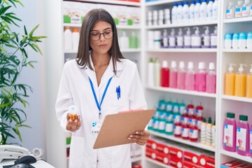 Young beautiful hispanic woman pharmacist holding pills bottle reading document at pharmacy