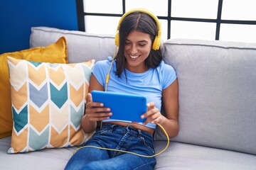 Young beautiful hispanic woman watching video on touchpad sitting on sofa at home
