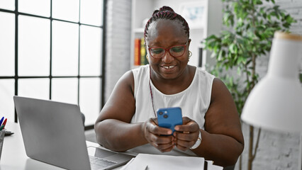 Confident african american business woman, working with laptop and smartphone, smiling boldly at the office interior
