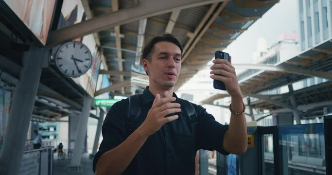 Male tourist with backpack making video call with smartphone while waiting for overground rail at city. Male tourist talking phone while waiting sky train at platform station of public transport