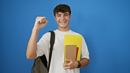 Happy young hispanic teenager student with backpack celebrating while holding books, standing isolated on a blue background in a cool, confident and smart lifestyle portrait.