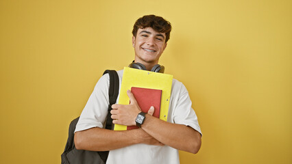 Cool young hispanic teenager boy wearing headphones, confidently holding books, smiling with joy...