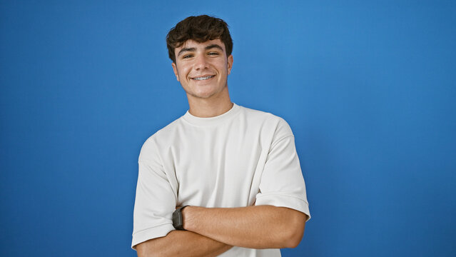 Joyous, confident young hispanic teenager guy standing with crossed arms, spreading positive vibes, cheerfully smiling against isolated blue background.