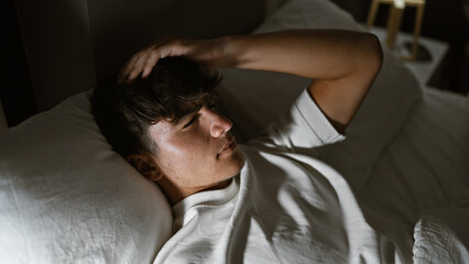 Handsome, young hispanic teenager lying comfortably in bed, engrossed in deep thought under a cozy...