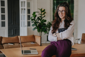 Brunette hispanic businesswoman in glasses sitting on desk with laptop smiles wide looks aside at break of work at office. Successful people. Happy accountant remote working home.