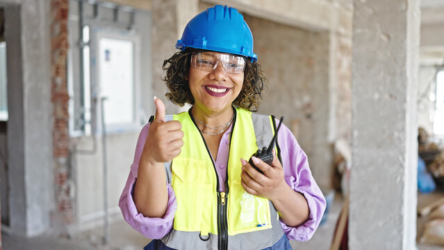 Young beautiful latin woman builder holding walkie talkie doing thumb up gesture at construction site