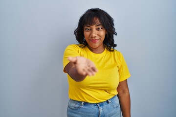 Hispanic woman standing over blue background smiling cheerful offering palm hand giving assistance and acceptance.