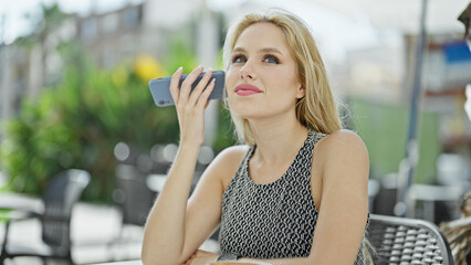 Young blonde woman listening to voice message by smartphone sitting on table at coffee shop terrace