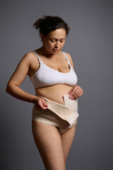 Portrait of naked woman in underwear after c-section, wearing elastic bandage, isolated over gray...