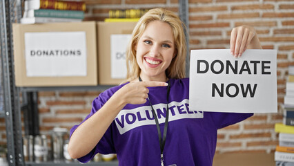 Young blonde woman volunteer smiling confident pointing to donate now banner at charity center