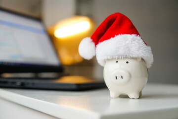 Piggy bank with red Santa hat and laptop on table. Christmas shopping and budgeting.