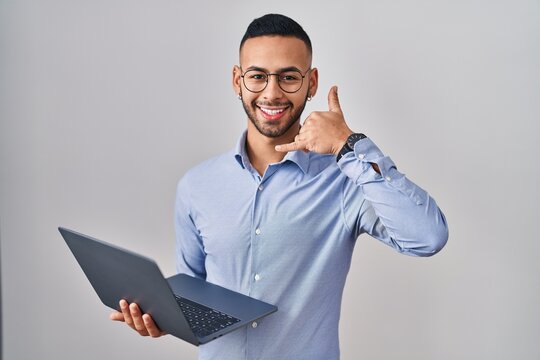 Young hispanic man working using computer laptop smiling doing phone gesture with hand and fingers like talking on the telephone. communicating concepts.