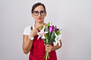 Middle age brunette woman wearing apron working at florist shop holding bouquet pointing aside...
