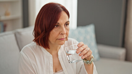 Middle age woman drinking glass of water sitting on sofa at home