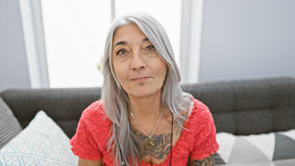 Captivating indoor shot of a beautiful, grey-haired middle age woman, deeply engrossed in relaxation, sitting comfortably in her cozy living room on a snug sofa with a serious, concentrated expression