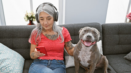 Grey-haired middle age woman sitting on living room sofa, absorbing her song's melody via gadget,...