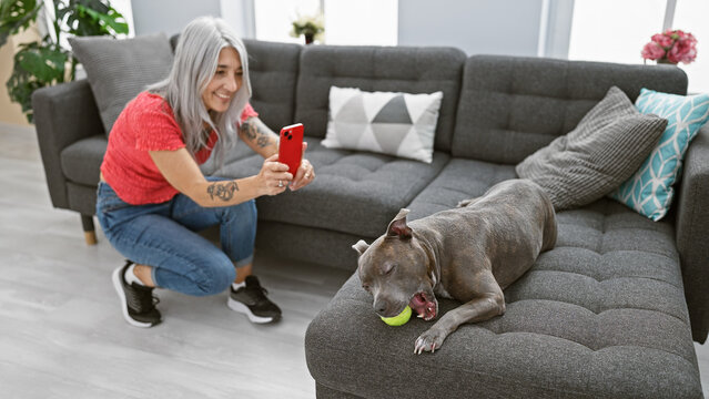 Joyful grey-haired middle age woman enjoys taking a pet dog picture with her smartphone while sitting on the homely indoor living room's sofa