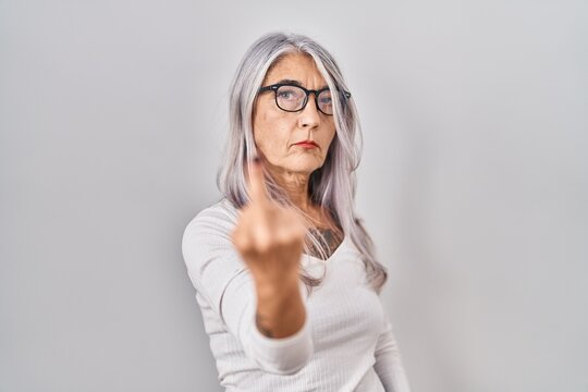Middle age woman with grey hair standing over white background showing middle finger, impolite and rude fuck off expression