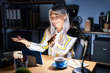 Middle age woman with grey hair working at the office at night looking at the camera smiling with open arms for hug. cheerful expression embracing happiness.