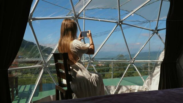 Woman captures mountain view from dome accommodation, showcasing modern travel lifestyle in nature.
