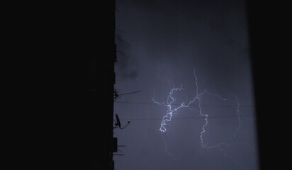 Night thunderstorm with lightning over the town and houses. In the foreground, the wall of a house with satellite dishes and air conditioners. Concept of weather, disasters and storms.