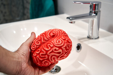 A man's hand holds a red rubber brain over a sink with a faucet, to perform brainwashing.