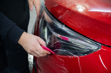 The master applies vinyl film to the headlight of a red car. Closeup view on worker detailer hand...