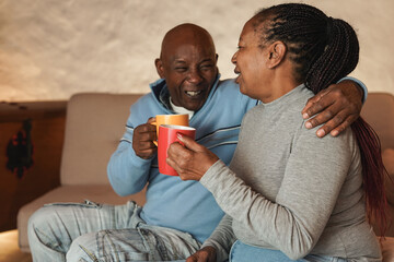 Happy african senior couple having tender moment together at home during winter time while drinking...