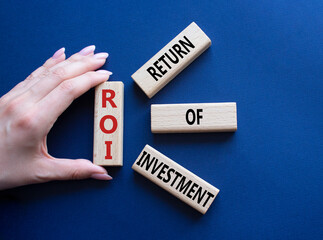 ROI - Return of Investment. Wooden cubes with words ROI. Businessman hand. Beautiful deep blue background. Business and ROI concept. Copy space.