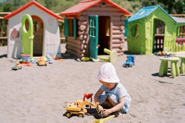 Little girl digs sand with a toy excavator while squatting