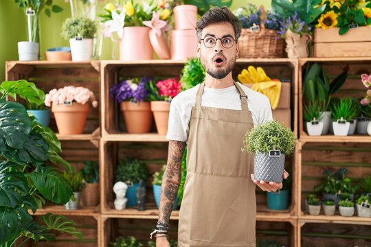 Young hispanic man with tattoos working at florist shop holding plant scared and amazed with open mouth for surprise, disbelief face